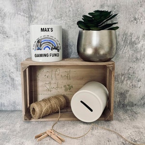 May include: White ceramic mug with a blue and gray rainbow design, a cartoon gamer holding a controller, and the text "MAX'S GAMING FUND". The mug is sitting on a wooden crate with a white ceramic piggy bank, twine, and a small wooden clothespin.