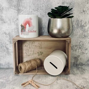 May include: Two white ceramic money boxes with a pink and gold rainbow design and the name Sadie on one. The money boxes are in a wooden crate with a sign that says "Kent Craft Studio Personalisation & Print".