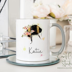 May include: White ceramic mug with a gray handle. The mug features a watercolor illustration of a bumblebee with yellow and black stripes surrounded by pink and yellow flowers. The name "Katie" is written in black cursive below the illustration.
