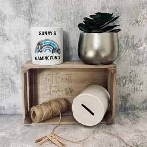 May include: White ceramic piggy bank with a slot on the top. The piggy bank is sitting in a wooden crate with a sign that says "Kent Craft Studio Personalisation & Print". There is a white ceramic mug with a blue and gray rainbow, a video game controller, and the text "Sonny's Gaming Fund" on it.