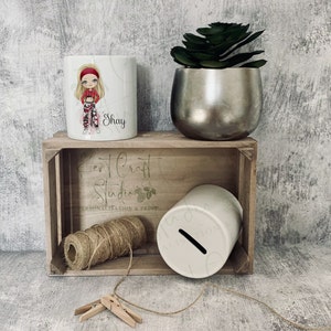 May include: White ceramic mug featuring a cartoon girl in a red and black plaid outfit and a red headband. The mug is personalised with the name "Shay". The mug is displayed on a wooden crate with a white ceramic money box and a ball of twine.