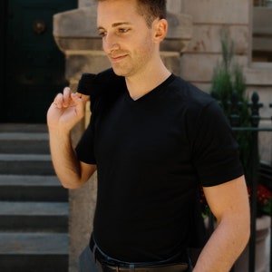 May include: A man wearing a black v-neck t-shirt, gray pants, and a silver watch. He is holding a black jacket over his shoulder.