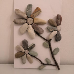 K&ouml;nnte beinhalten: Ein wei&szlig;er Hintergrund mit zwei braunen Zweigzweigen mit drei kleinen gr&uuml;nen Bl&auml;ttern und zwei Blumen aus verschiedenen farbigen Steinen. Die Blumen haben die Form von G&auml;nsebl&uuml;mchen mit einer Mitte aus einem kleinen runden Stein.