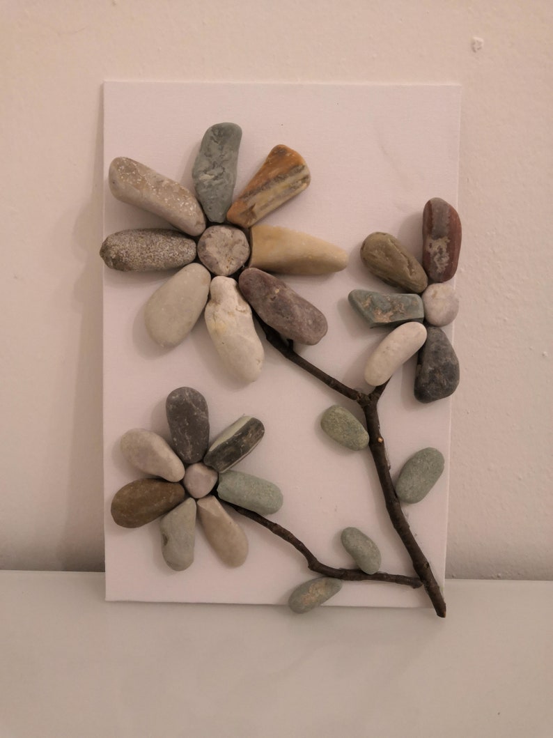 K&ouml;nnte beinhalten: Ein wei&szlig;er Hintergrund mit zwei braunen Zweigzweigen mit drei kleinen gr&uuml;nen Bl&auml;ttern und zwei Blumen aus verschiedenen farbigen Steinen. Die Blumen haben die Form von G&auml;nsebl&uuml;mchen mit einer Mitte aus einem kleinen runden Stein.