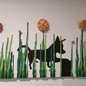 May include: A black silhouette of a dog walking through a field of green crayon art. The dog is walking towards a small flower. There are three orange flowers at the top of the art piece.