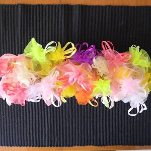 K&ouml;nnte beinhalten: Eine Sammlung von bunten, durchsichtigen Stoffbeuteln mit Kordeln. Die Beutel sind in den Farben Rosa, Gelb, Orange, Gr&uuml;n und Lila. Die Beutel sind in einem Halbkreis auf einer schwarzen, strukturierten Oberfl&auml;che angeordnet.