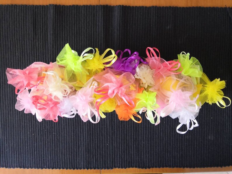 K&ouml;nnte beinhalten: Eine Sammlung von bunten, durchsichtigen Stoffbeuteln mit Kordeln. Die Beutel sind in den Farben Rosa, Gelb, Orange, Gr&uuml;n und Lila. Die Beutel sind in einem Halbkreis auf einer schwarzen, strukturierten Oberfl&auml;che angeordnet.