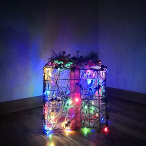 May include: A Christmas tree shaped like a gift box, wrapped in twine and decorated with colourful lights. The tree is lit up with multi-coloured lights, creating a festive atmosphere.
