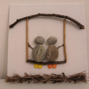 K&ouml;nnte beinhalten: Ein Steinbild, das zwei Figuren auf einer Schaukel aus Zweigen und Schnur zeigt. Die Figuren sind aus grauen Steinen mit gelben und orangefarbenen Akzenten f&uuml;r ihre Beine.