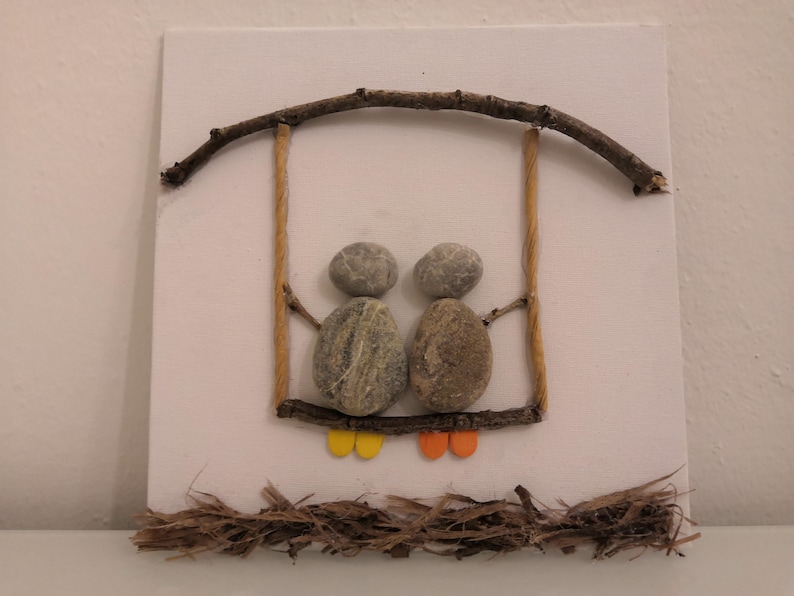 K&ouml;nnte beinhalten: Ein Steinbild, das zwei Figuren auf einer Schaukel aus Zweigen und Schnur zeigt. Die Figuren sind aus grauen Steinen mit gelben und orangefarbenen Akzenten f&uuml;r ihre Beine.