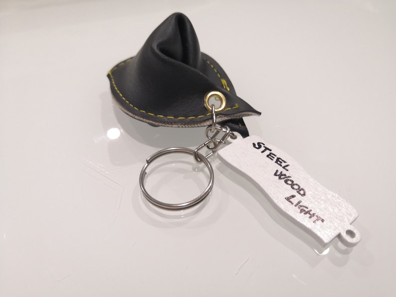 K&ouml;nnte beinhalten: Schwarzer Leder-Schl&uuml;sselanh&auml;nger mit gelber Naht und einem wei&szlig;en Etikett mit der Aufschrift "STEEL WOOD LIGHT".