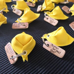 K&ouml;nnte beinhalten: Nahaufnahme von gelben Stoff-Tortellini-Schl&uuml;sselanh&auml;ngern mit Holzetiketten, auf denen "Teatro Sociale 100" und "E Capulet" steht.