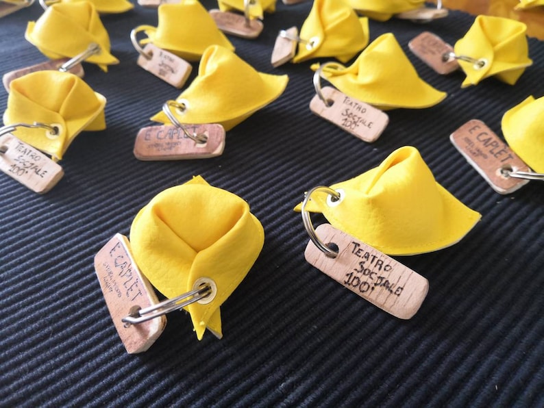 K&ouml;nnte beinhalten: Nahaufnahme von gelben Stoff-Tortellini-Schl&uuml;sselanh&auml;ngern mit Holzetiketten, auf denen "Teatro Sociale 100" und "E Capulet" steht.