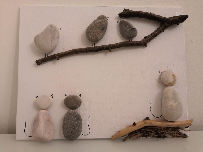 K&ouml;nnte beinhalten: Eine wei&szlig;e Leinwand mit einem Ast, auf dem drei graue V&ouml;gel und zwei graue und wei&szlig;e Katzen aus Steinen sitzen. Die Katzen sitzen unten auf der Leinwand und die V&ouml;gel sitzen auf dem Ast.