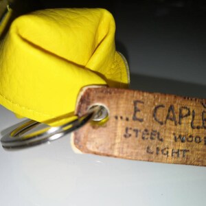 K&ouml;nnte beinhalten: Ein gelber Leder-Schl&uuml;sselanh&auml;nger mit einem Holzschild, auf dem "...E CAPLET... STEEL WOOD LIGHT" steht.