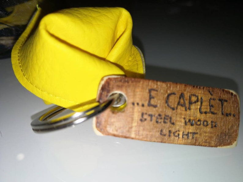 K&ouml;nnte beinhalten: Ein gelber Leder-Schl&uuml;sselanh&auml;nger mit einem Holzschild, auf dem "...E CAPLET... STEEL WOOD LIGHT" steht.