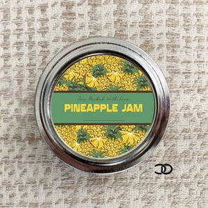May include: A silver jar lid with a green and yellow label that says "Home Picked With Love PINEAPPLE JAM". The label features a pattern of pineapple slices.