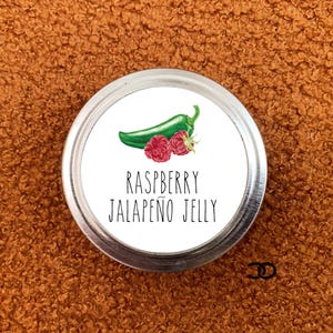 May include: A silver jar with a white label that reads "Raspberry Jalapeño Jelly". The label features a green jalapeño pepper and two red raspberries.