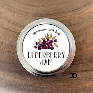 May include: White round jar lid with a white label that says "homemade with love" and features a watercolor illustration of elderberries. The label also says "Elderberry Jam".