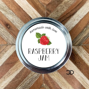 May include: White round jar lid with a white label that says "homemade with love" and "raspberry jam" in black text. A red raspberry with green leaves is pictured on the label.
