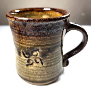 Puede incluir: Taza de cerámica hecha a mano con un esmalte marrón y beige. La taza tiene un asa marrón oscuro y un símbolo decorativo cerca de la base. El exterior de la taza tiene estrías horizontales. Adecuada para bebidas frías o calientes.