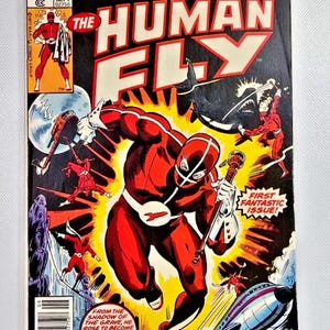Puede incluir: Una portada de cómic vintage con "The Human Fly" con un traje rojo y blanco, empuñando un bastón. La portada tiene un fondo negro con explosiones amarillas y escenas de acción. El texto incluye "Marvel Comics Group" y "First Fantastic Issue!"