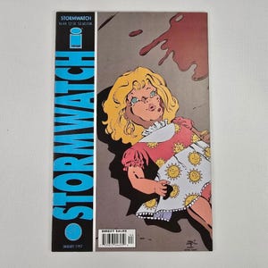 Puede incluir: Portada del cómic Stormwatch con un personaje parecido a una muñeca, con cabello rubio y un vestido rojo con diseños de sol. El título "Stormwatch" está en letras azules. La portada tiene un tono oscuro y dramático.
