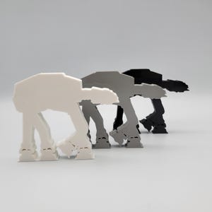 Pode incluir: Três estatuetas de caminhantes AT-AT em branco, cinza e preto. Os modelos inspirados em Star Wars são exibidos em um fundo cinza claro. As estatuetas são estilizadas e têm quatro pernas.
