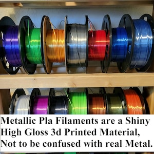 Op de afbeelding: Een plank met spoelen van metallic PLA-filament voor 3D-printen. Het filament is in verschillende kleuren verkrijgbaar, waaronder paars, groen, oranje, zilver, rood, blauw, wit, roze, zwart, limoengroen, koper en bruin. De tekst op de plank luidt "Metallic Pla Filaments are a Shiny High Gloss 3d Printed Material, Not to be confused with real Metal."