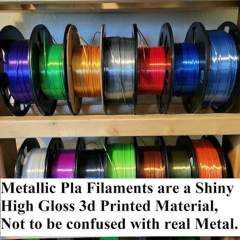Op de afbeelding: Een plank met spoelen van metallic PLA-filament voor 3D-printen. Het filament is in verschillende kleuren verkrijgbaar, waaronder paars, groen, oranje, zilver, rood, blauw, wit, roze, zwart, limoengroen, koper en bruin. De tekst op de plank luidt "Metallic Pla Filaments are a Shiny High Gloss 3d Printed Material, Not to be confused with real Metal."