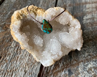 May include: A turquoise teardrop pendant on a brown cord necklace. The pendant is resting on a white crystal geode.