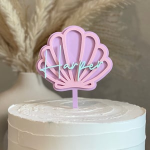 May include: A white cake with pink and green frosting and a pink seashell topper with the name "Harper" written on it.