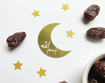 May include: A gold crescent moon with the Arabic phrase "Bismillah" written inside, surrounded by small gold stars. Dates are scattered around the moon and in a bowl.