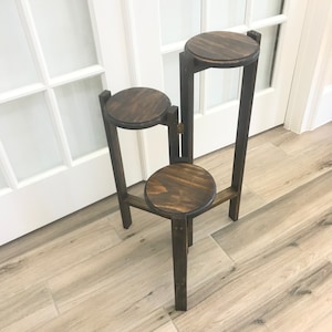 May include: A dark brown, three-tiered plant stand made of wood. The stand has three circular platforms of varying heights, connected by vertical supports. The wood has a visible grain and a stained finish. The stand is designed to hold plants.