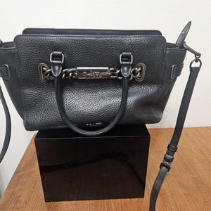 May include: Black leather handbag with a top handle and a detachable shoulder strap. The bag features silver-tone hardware, including a chain detail across the front. The bag is sitting on a black box.