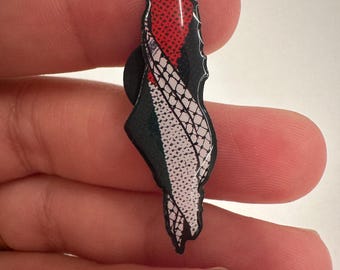 May include: A pin shaped like the outline of Israel with the Israeli flag colors, red, white, and blue, and a black and white snake skin pattern.