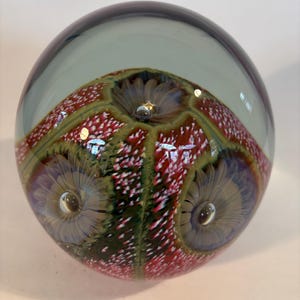 May include: A spherical glass paperweight featuring a clear top section and an internal design of red, white, and green speckled patterns. The core contains three distinct, circular, flower-like motifs with radiating petals and central air bubbles.