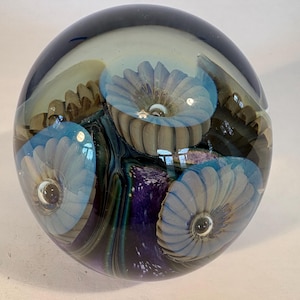 May include: A spherical glass paperweight featuring three embedded light blue flower-like designs with ribbed petals. The interior contains deep purple and dark blue swirling patterns with subtle white speckles encased in clear glass.