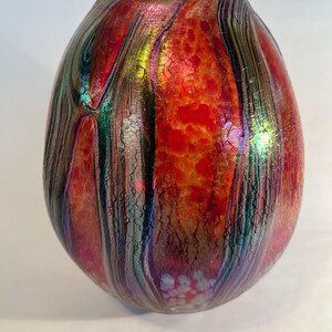 May include: A red, green, and iridescent glass vase with a textured surface. The vase has a rounded, egg-like shape.