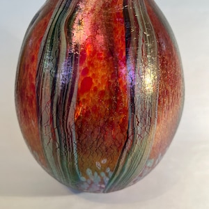May include: A round, iridescent glass vase with a textured surface. The vase has a mix of red, orange, green, and silver colors.