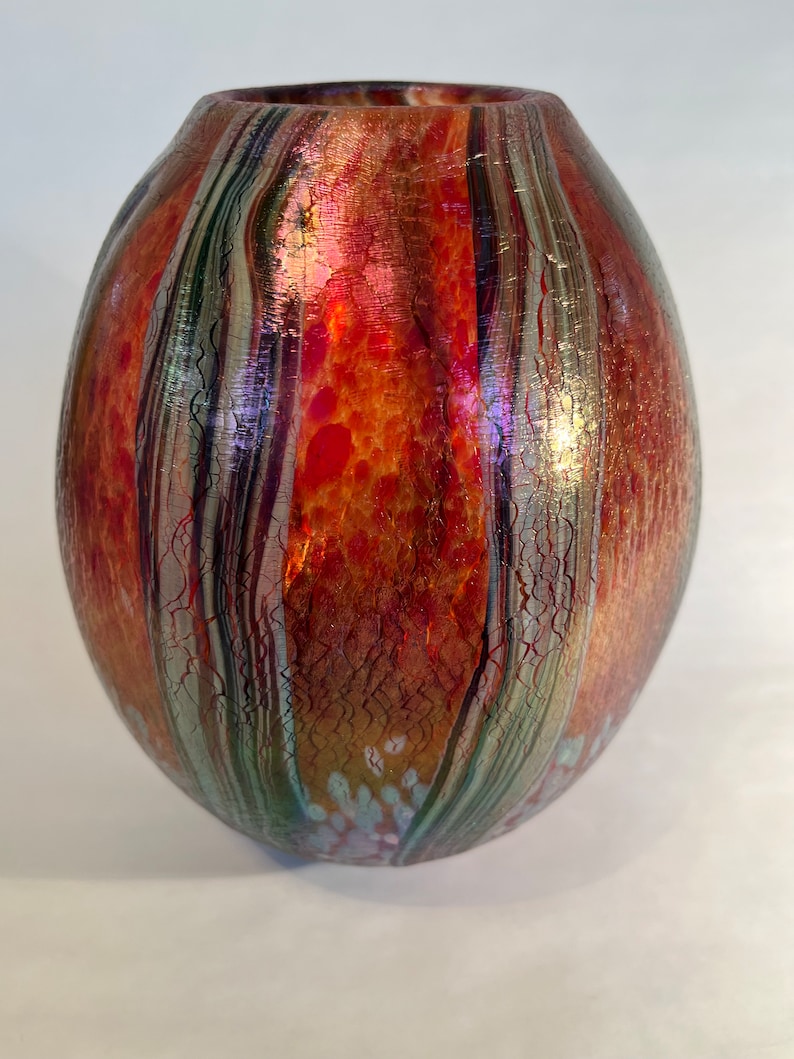 May include: A round, iridescent glass vase with a textured surface. The vase has a mix of red, orange, green, and silver colors.
