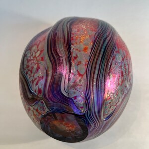May include: A round glass vase with a swirling pattern of purple, blue, and red iridescent glass. The vase has a textured surface with small white dots.