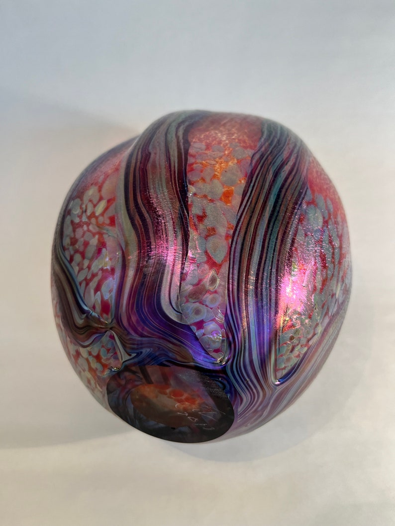 May include: A round glass vase with a swirling pattern of purple, blue, and red iridescent glass. The vase has a textured surface with small white dots.