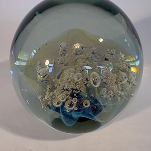 May include: A spherical glass paperweight featuring a clear, smoke-gray outer layer. The interior contains a cluster of suspended air bubbles and swirling accents of pale yellow and blue glass, creating a layered, multi-dimensional effect.