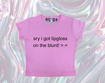 May include: A short-sleeved, pink t-shirt with the text "sry i got lipgloss on the blunt! >.<" in black. The shirt has a crew neck and rolled sleeves. The background is a swirling, iridescent pattern of pink, white, and silver.