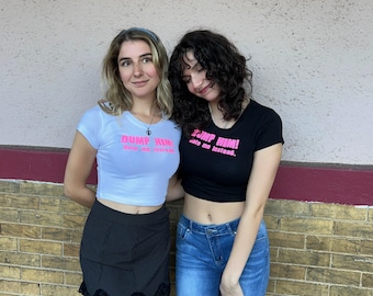 May include: Two people wearing crop tops with the text "Dump Him! Date me instead." printed on them. One person is wearing a white crop top with black pinstripe skirt and the other is wearing a black crop top with blue jeans.
