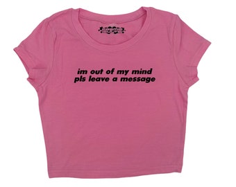 May include: A pink crop top with a crew neck and short sleeves. The text "im out of my mind pls leave a message" is printed in black across the front. The top has a small logo near the neckline.