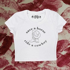 Op de afbeelding: Wit kort T-shirt met een zwarte afbeelding van cowboylaarzen en een hoed. De tekst "save a horse ride a cowboy" is op het shirt gedrukt.