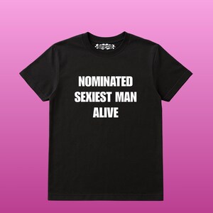 May include: Black crew neck t-shirt with the text "NOMINATED SEXIEST MAN ALIVE" in white block letters. The shirt has short sleeves and a small logo at the neckline. The background is a gradient of pink and purple.