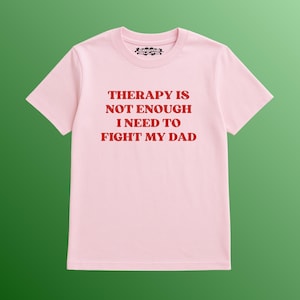 May include: A light pink t-shirt with the text "THERAPY IS NOT ENOUGH I NEED TO FIGHT MY DAD" in red block letters. The shirt has a crew neck and short sleeves. The shirt is a solid color with no other design elements.